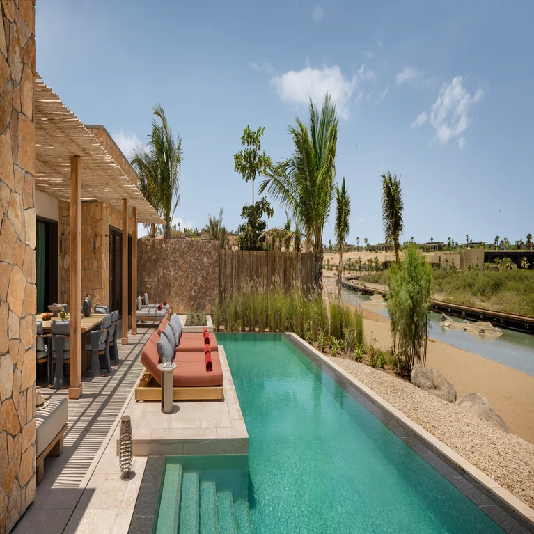 Villa featuring a private pool, sun loungers, and open-air dining space amid palm trees, with views of a sandy riverbank and clear sky.
