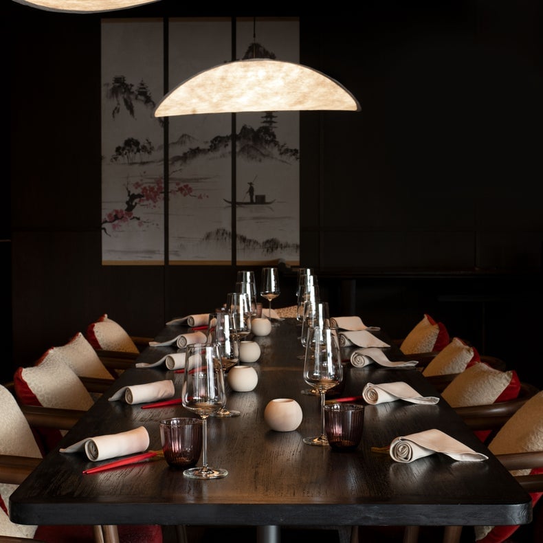 Salle à manger avec tables, chaises moelleuses, éclairage suspendu organique au-dessus et œuvres d'art d'inspiration asiatique sur un mur sombre pour une ambiance accueillante.