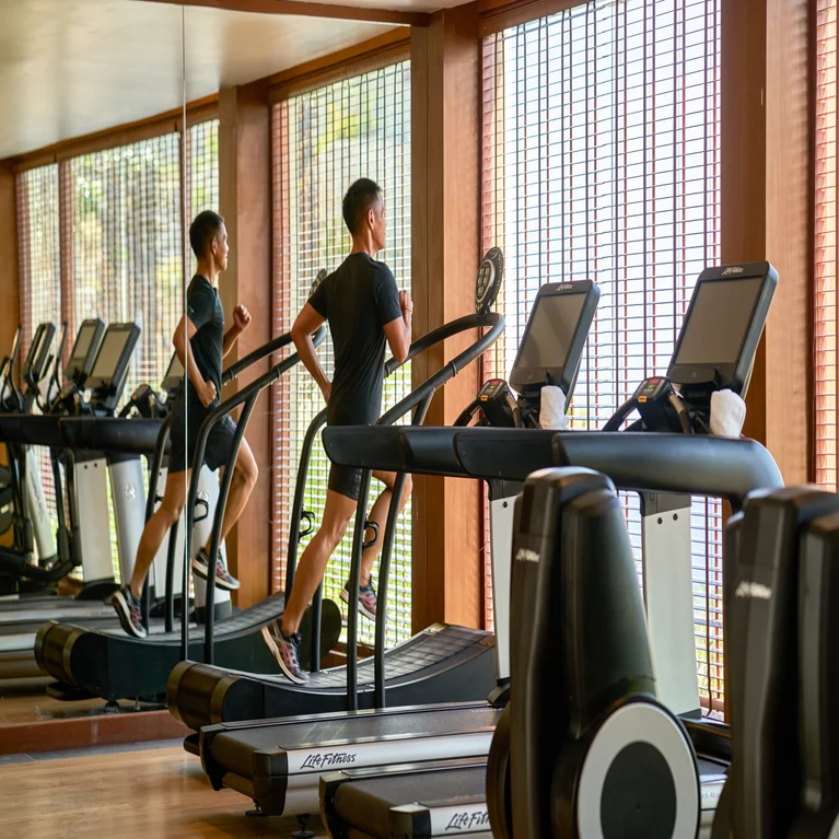 Une personne faisant du jogging sur un tapis de course dans une salle de sport baignée de lumière, dont l'image se reflète dans un miroir allant jusqu'au sol, à côté de grandes baies vitrées donnant sur un paysage naturel.