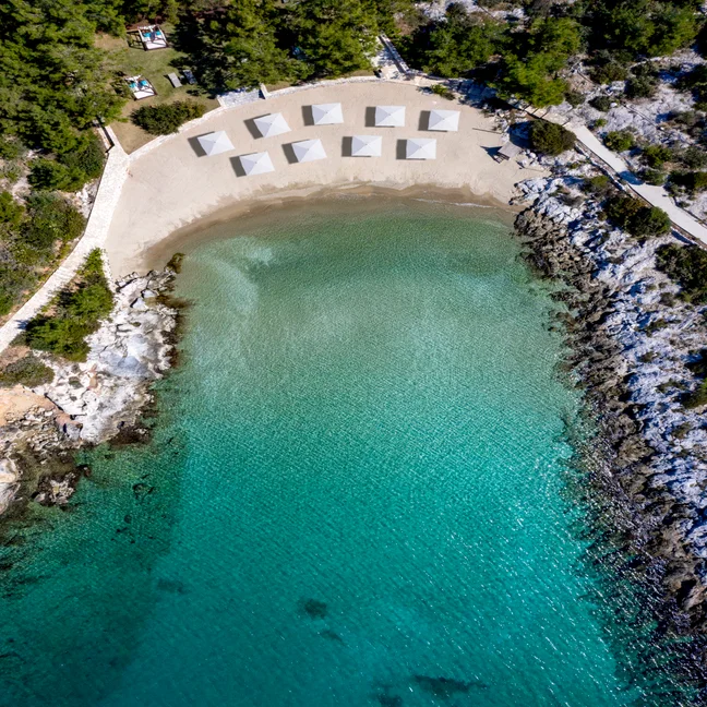 View of a serene sandy beach bordered by turquoise water, rocky outcrops, and pine trees, with evenly spaced white sunshades available for guests.