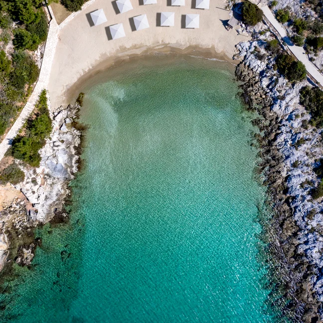 View of a serene sandy beach bordered by turquoise water, rocky outcrops, and pine trees, with evenly spaced white sunshades available for guests.