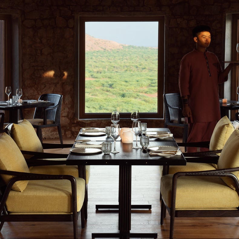 Comedor con ventanales con vistas a las verdes colinas, cuatro mesas espaciosas y un camarero con uniforme regional que lleva una bandeja.
