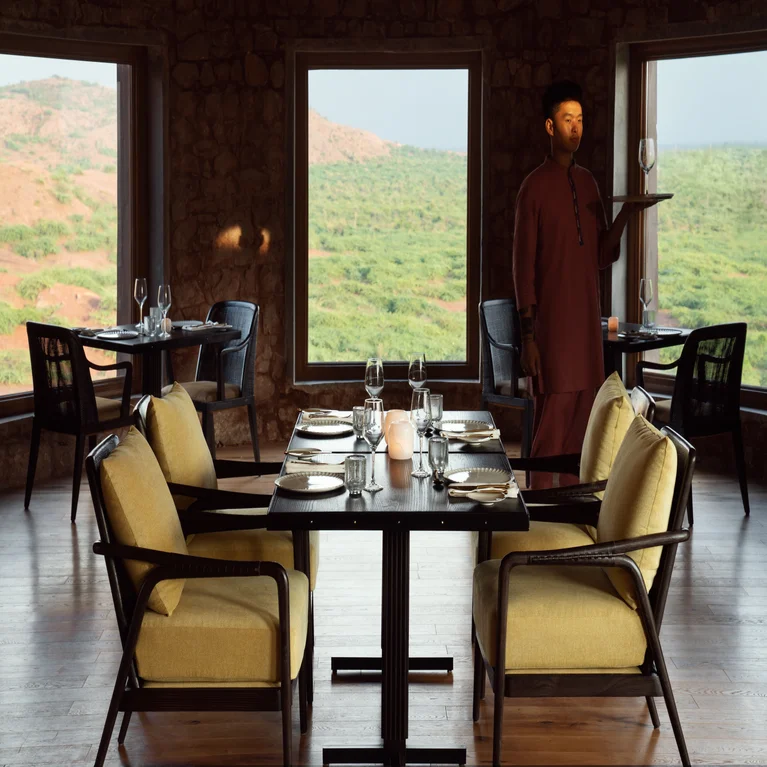 Comedor con ventanales con vistas a las verdes colinas, cuatro mesas espaciosas y un camarero con uniforme regional que lleva una bandeja.