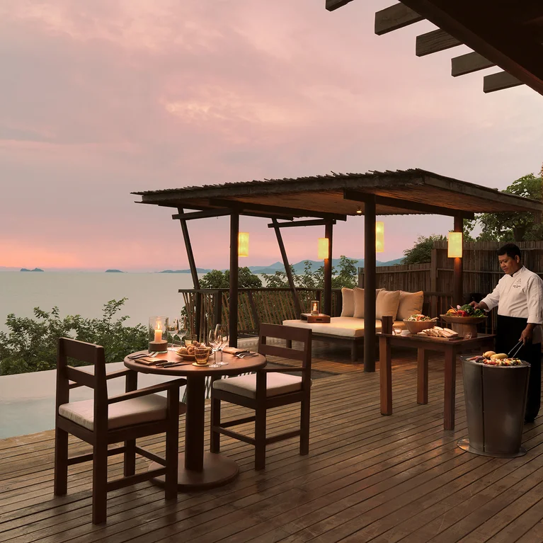 Dining tables prepared on a wooden terrace above the sea at sunset, with outdoor seating beneath pergolas providing a tranquil setting.