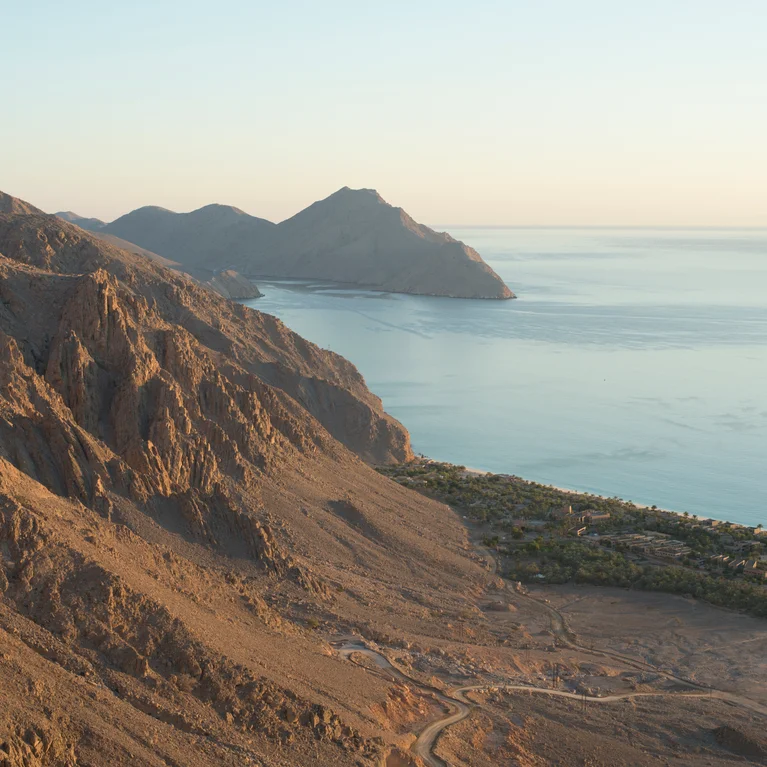 Las laderas arboladas se encuentran con tranquilas aguas azules, con un pico lejano y un pequeño pueblo cerca de la orilla, todo suavemente iluminado por la luz natural del día.