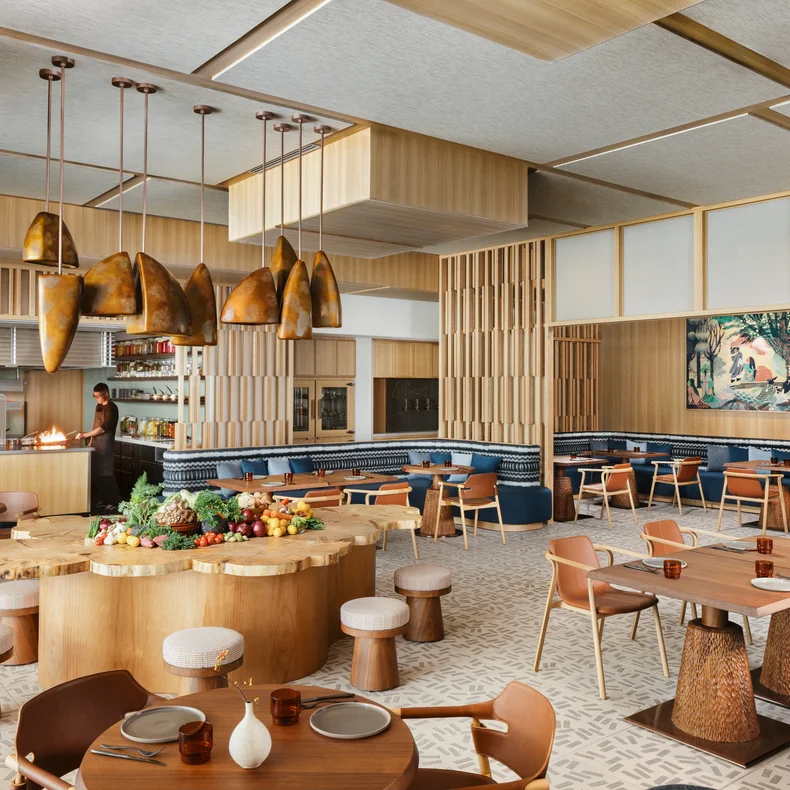 木製の椅子、木の幹のようなテーブル、ペンダントライト、青いクッションのベンチ、壁画のウォールアート、テーブルが配置されたレストラン。.