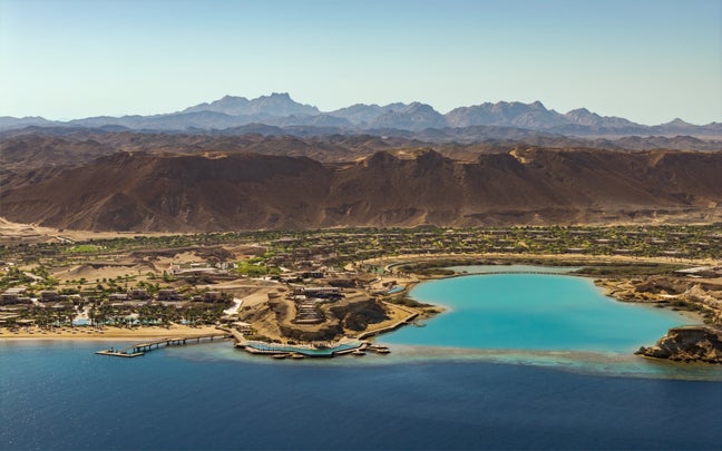 Coastal resort with turquoise lagoons, a wooden pier, desert mountains beyond, and blue sea in the foreground under clear skies.