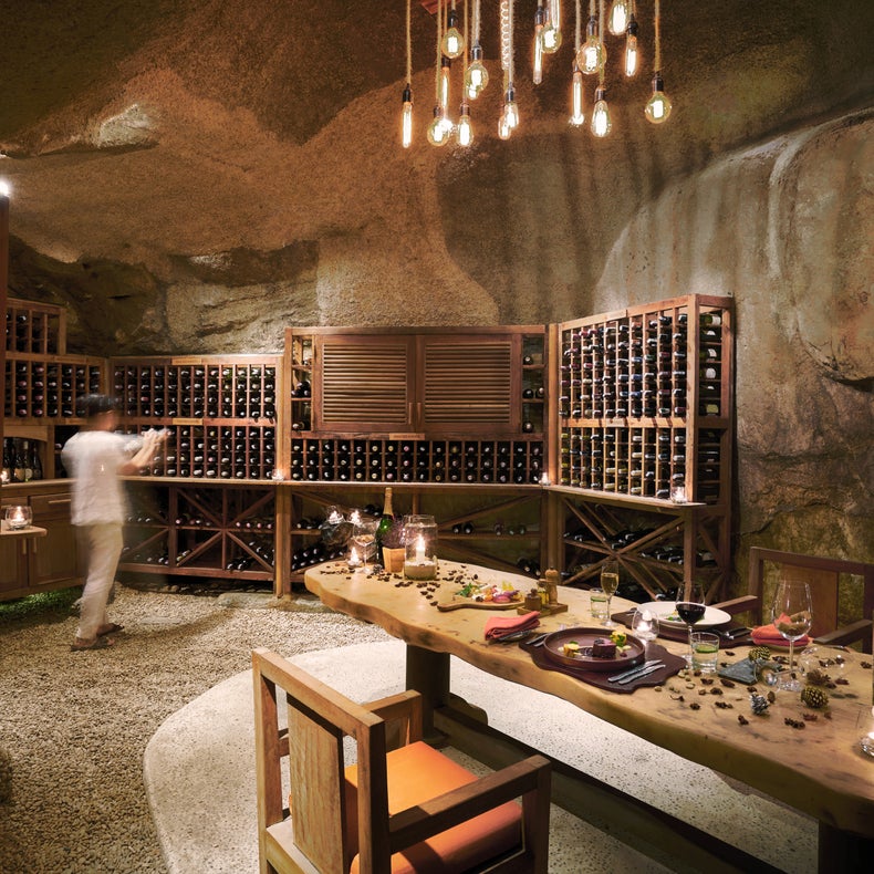 Une cave à vin dans une grotte avec des étagères en bois, une table dressée pour le repas et un éclairage tamisé ; une personne se tient à côté de l'étagère, légèrement floue.