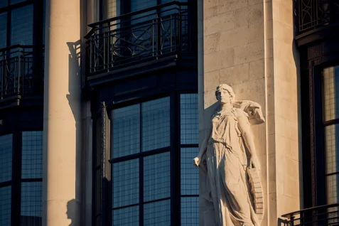 Stone statue of a robed figure stands in front of a beige building with tall windows and black metal balconies, emphasizing refined design and tranquil atmosphere.