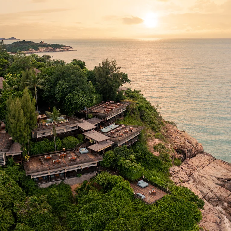 木制别墅和露台坐落在岩石海岸线上茂密的绿树丛中,在柔和的落日余晖下俯瞰宁静的海面。.