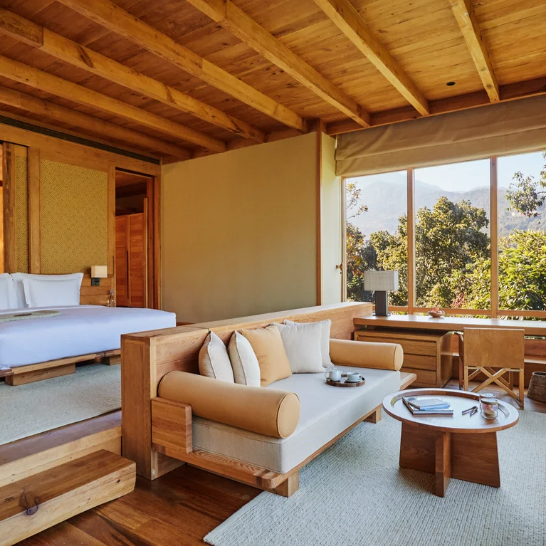 Bedroom features natural wood details, king bed, sofa, and coffee table. Floor-to-ceiling windows offer views of surrounding trees and distant mountains.