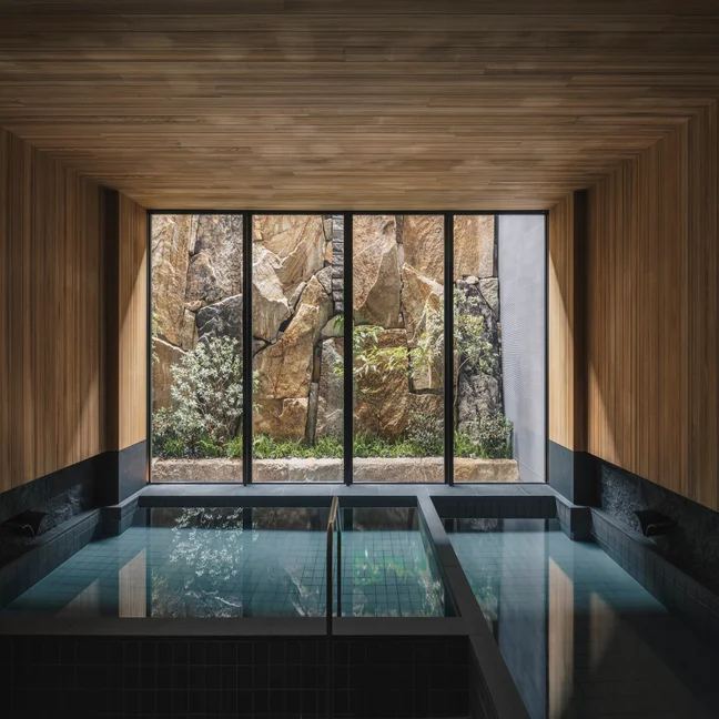 Tranquil swimming area bordered by natural wood accents and expansive windows that open to views of a serene garden with stones and lush plants.