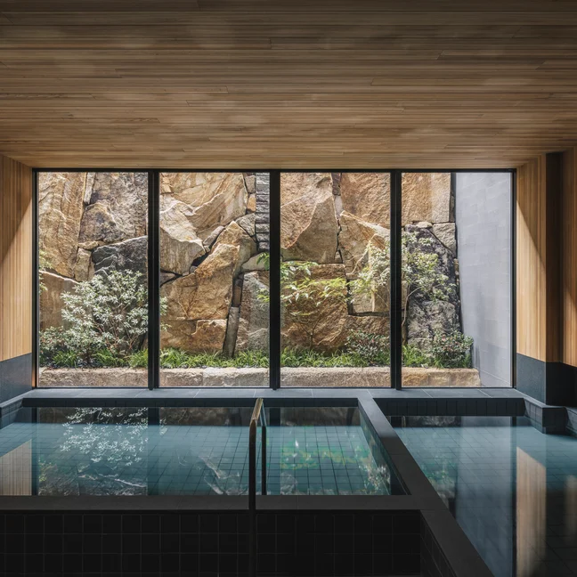Tranquil swimming area bordered by natural wood accents and expansive windows that open to views of a serene garden with stones and lush plants.