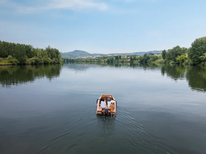 Two people in a wooden boat glide along a tranquil river. Green trees line the banks, with hills and a clear sky visible in the distance.