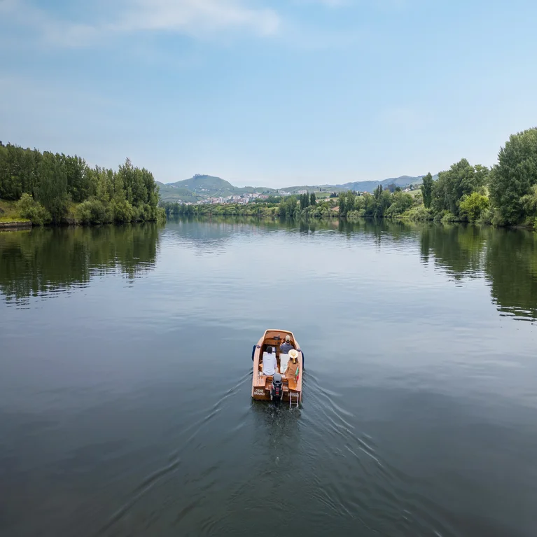 Two people in a wooden boat glide along a tranquil river. Green trees line the banks, with hills and a clear sky visible in the distance.