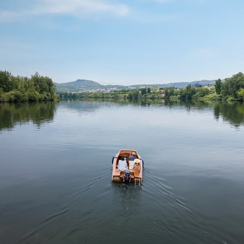 Two people in a wooden boat glide along a tranquil river. Green trees line the banks, with hills and a clear sky visible in the distance.