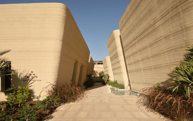 Stone path bordered by tall, curved beige walls, with natural greenery and a small water feature beneath a clear blue sky.