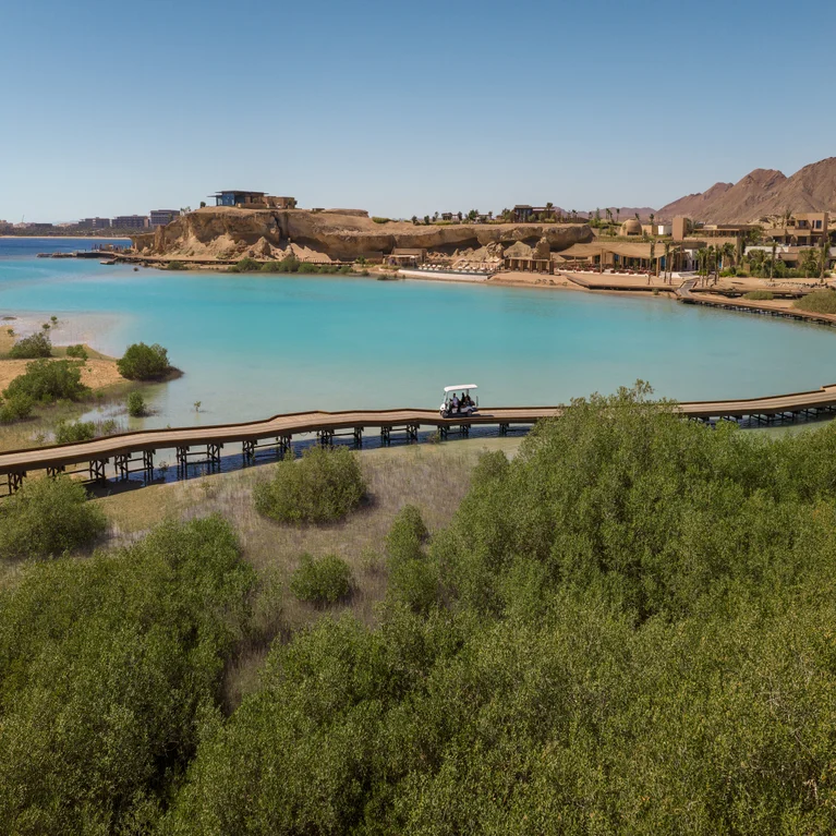 Electric cart drives across a wooden bridge surrounded by greenery, with turquoise water, sandy hills, and buildings in the distance under a clear sky.
