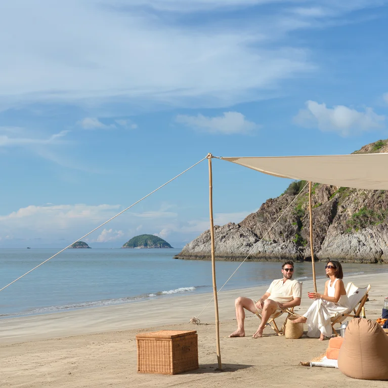 两位客人在沙滩上的顶篷下放松身心,在晴朗的天空下,面对平静的海水和岛屿,摆放着豆袋和野餐必需品。.