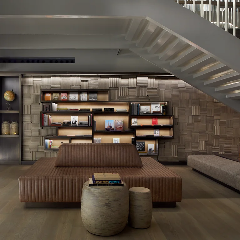 El tranquilo salón está amueblado con un sofá en tonos naturales, taburetes de madera, estanterías accesibles, iluminación con velas y una escalera de barandilla blanca....