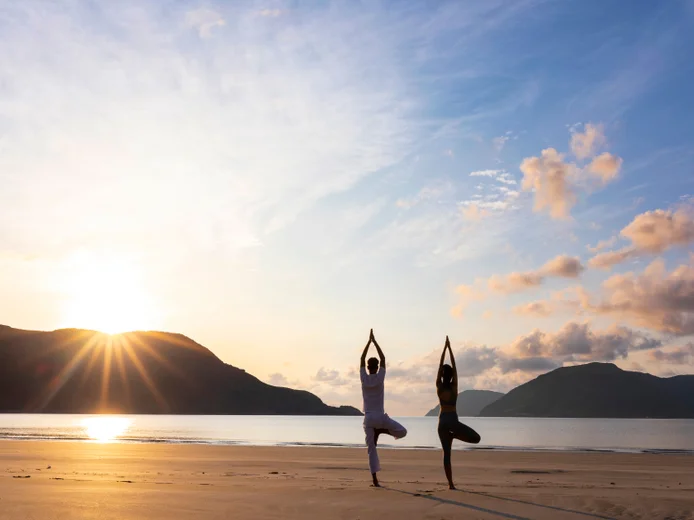Morning yoga on a sandy beach, framed by calm sea and distant mountains under soft sunrise light, inviting reflection and connection to nature.