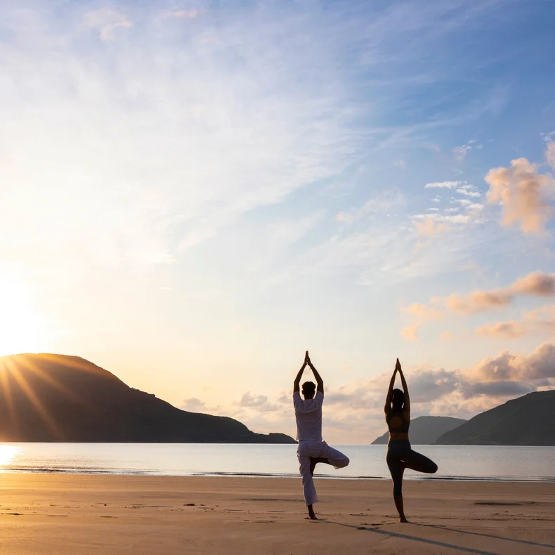 Morning yoga on a sandy beach, framed by calm sea and distant mountains under soft sunrise light, inviting reflection and connection to nature.