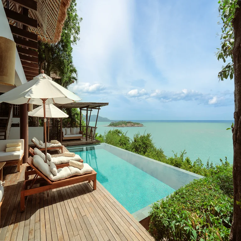 Serene pool with shaded lounge chairs on a wooden deck, overlooking green foliage and clear turquoise sea beneath a bright blue sky.
