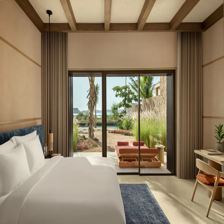 Spacious guest room with king bed and work area, featuring glass doors leading to a patio overlooking lush plants and distant ocean views, designed for comfort and relaxation.