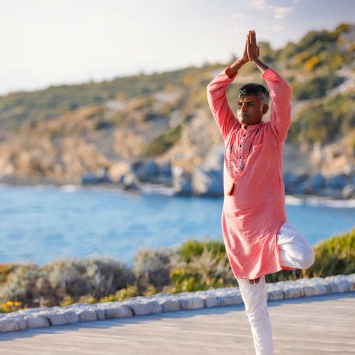 Person holding tree pose on a yoga mat by the water, with rocky hills in the background and a partly cloudy sky, highlighting balance and natural surroundings.