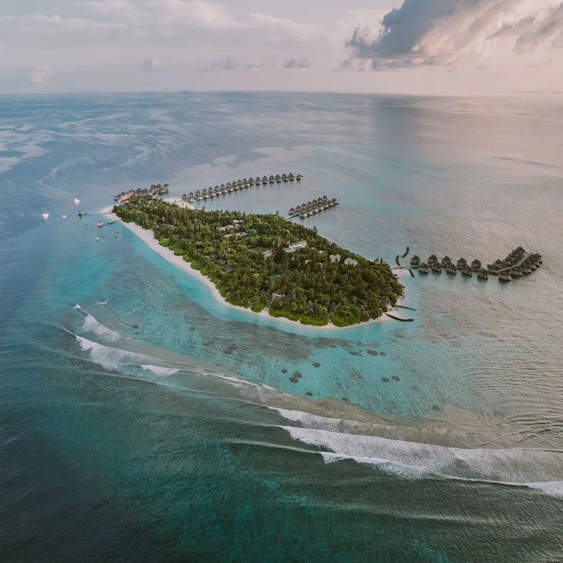 Overwater villas rest above clear blue water and white sand, blending into verdant island greenery beneath a sky with soft clouds.