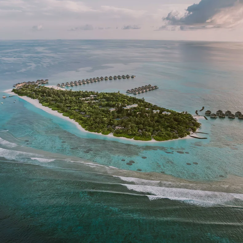 Overwater villas rest above clear blue water and white sand, blending into verdant island greenery beneath a sky with soft clouds.