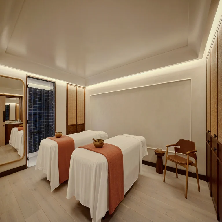 Spa room featuring two massage tables with white linens and orange accents, wooden bowls, a chair, and gentle ambient lighting.