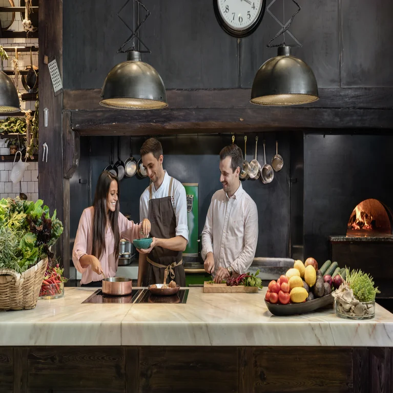 Guests prepare healthy dishes at a marble kitchen counter with fresh vegetables, illuminated by pendant lights, with a wood-fired oven and clock in the background.