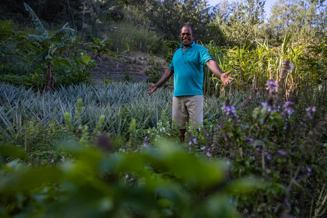 A man in a teal shirt stands with open arms in a vibrant garden, surrounded by diverse plants and crops that highlight natural abundance and wellness.