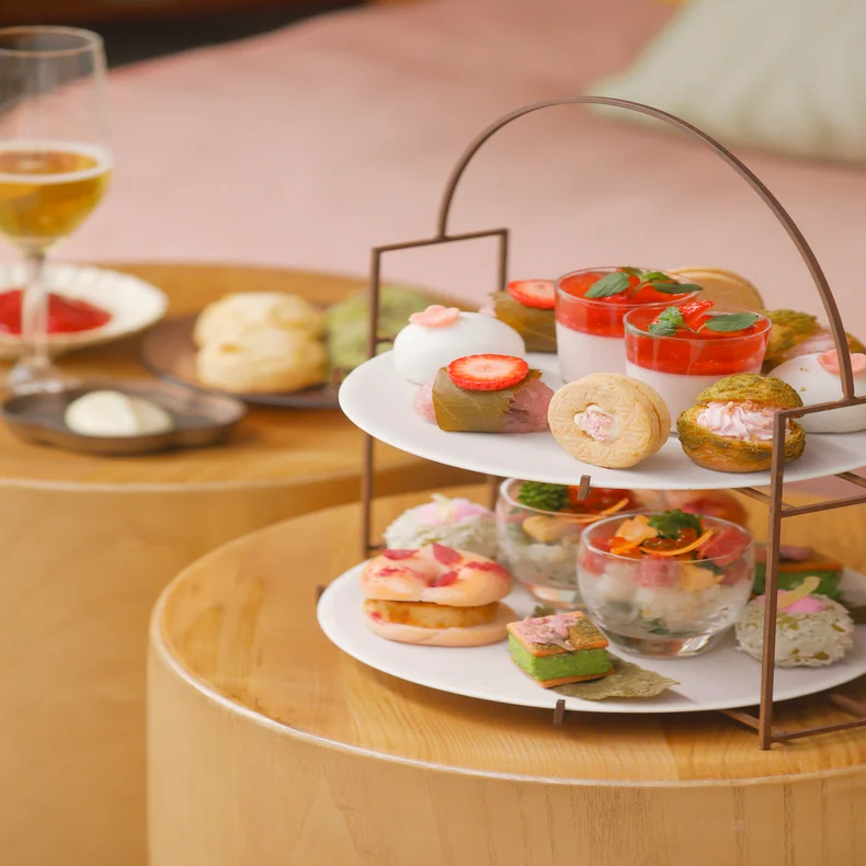 ペストリー、デザート、サンドイッチの盛り合わせが、シャンパンの横に2段トレイで並べられ、テーブルの上にはスコーン、クリーム、ジャムが乗ったプレートが置かれている。.