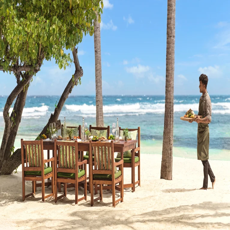 توجد طاولة طعام بستة كراسي على الرمال تحت أشجار النخيل، وتطل على المحيط. يجلب أحد الموظفين الطعام لتعزيز تجربة الاسترخاء على شاطئ البحر.