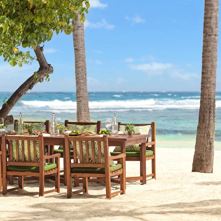 توجد طاولة طعام بستة كراسي على الرمال تحت أشجار النخيل، وتطل على المحيط. يجلب أحد الموظفين الطعام لتعزيز تجربة الاسترخاء على شاطئ البحر.