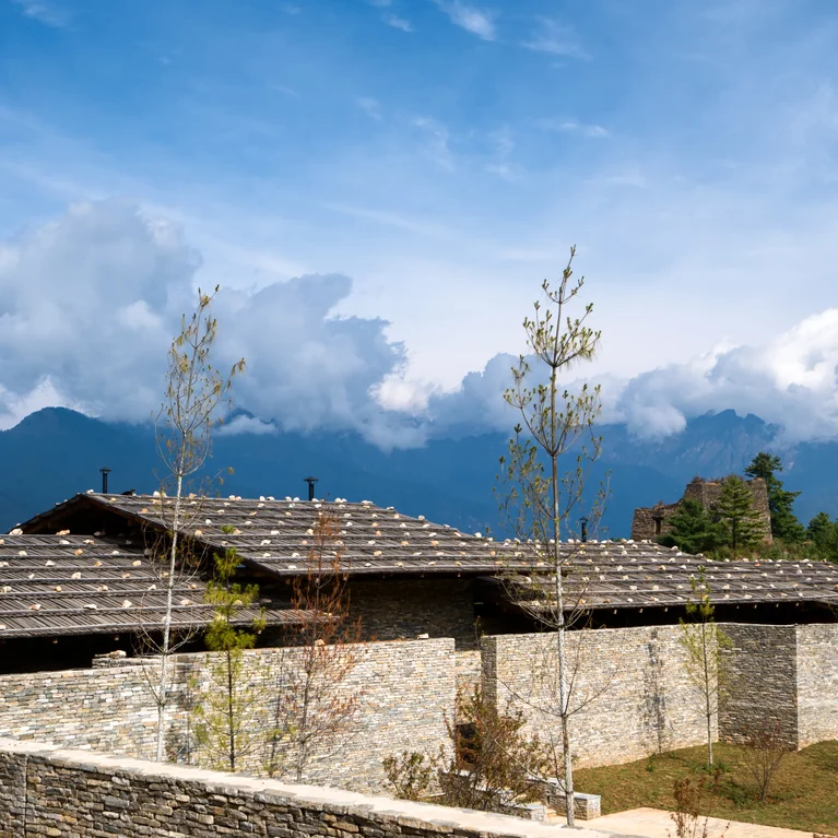 Stone building with a tiled roof beside a stone wall, framed by trees and mountains beneath a blue sky with soft clouds.