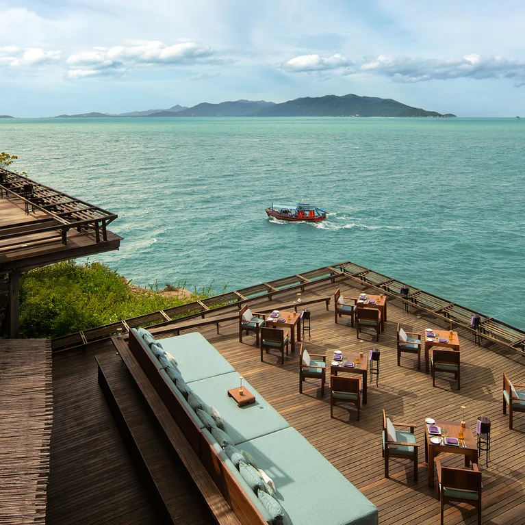 摆放着餐桌的露台面朝清澈蔚蓝的大海,一艘小船从海面上驶过。远处群山耸立,天空中云雾缭绕。.