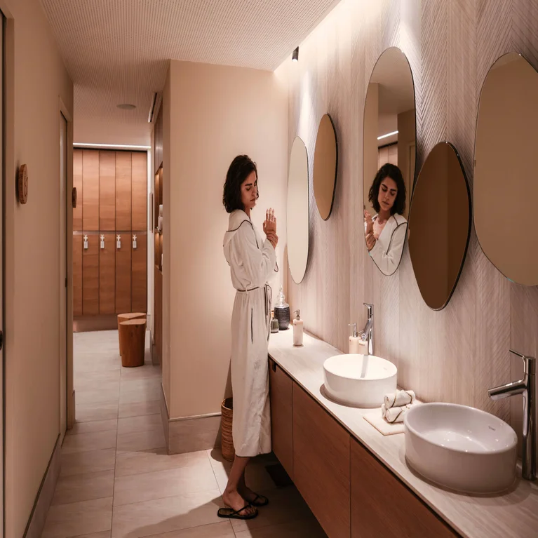 A guest in a white bathrobe applies lotion to their hands at a vanity with circular mirrors in a spa washroom.