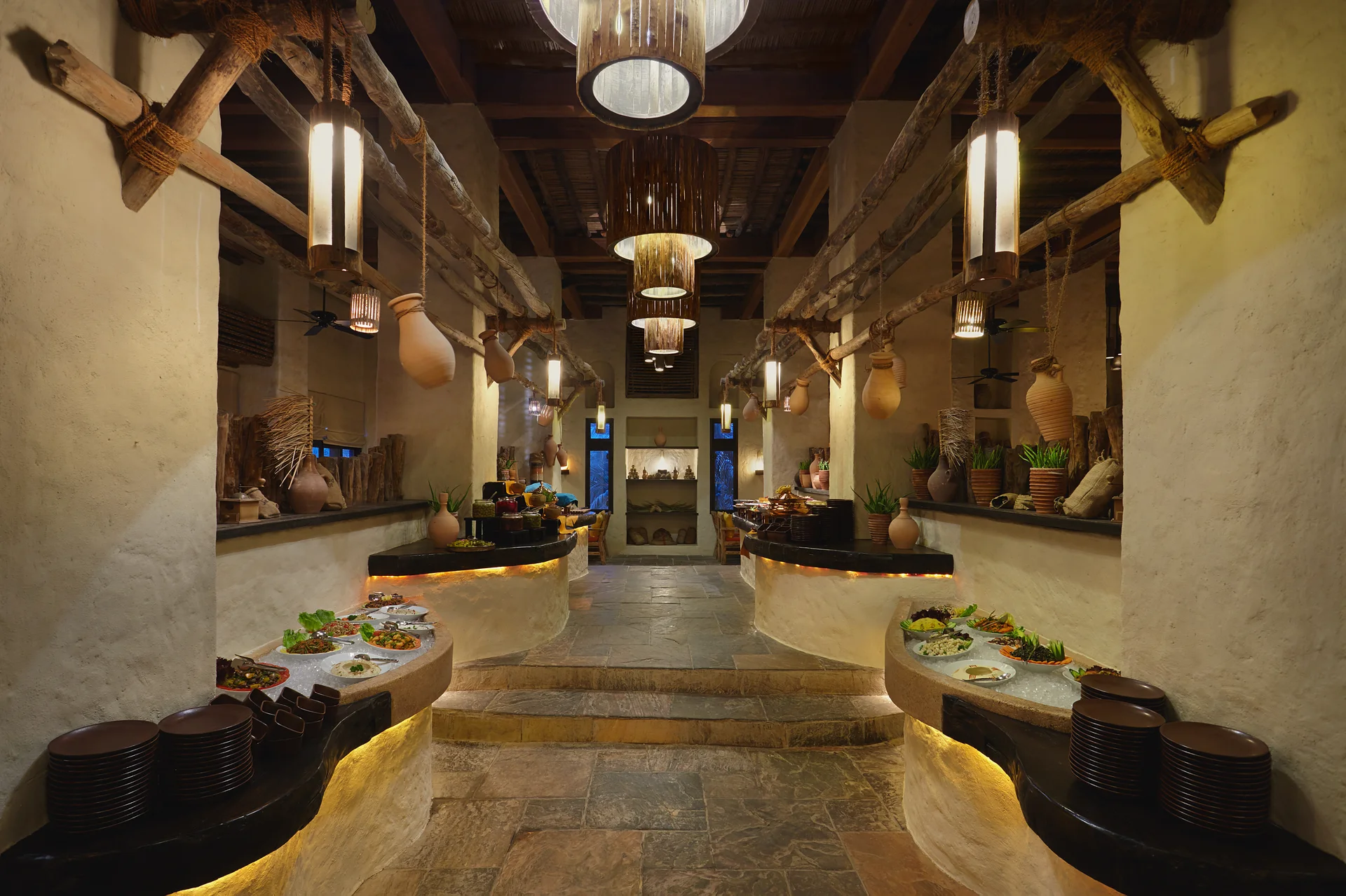 Spice Market at night, a long stone-floored hall with rough timber beams and hanging fibre lanterns, curved buffet counters of laid dishes lit from beneath and clay storage jars ranged along the walls.