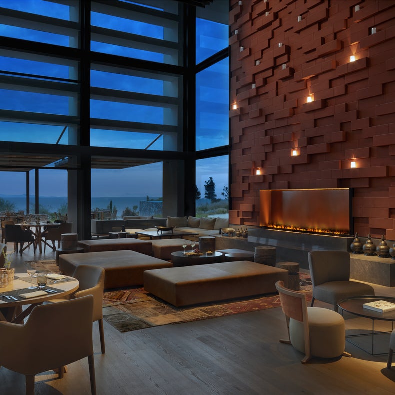 Un restaurante con materiales naturales, iluminación cálida, una acogedora chimenea y una amplia vista de la terraza y el paisaje nocturno gracias a los ventanales que van del suelo al techo.