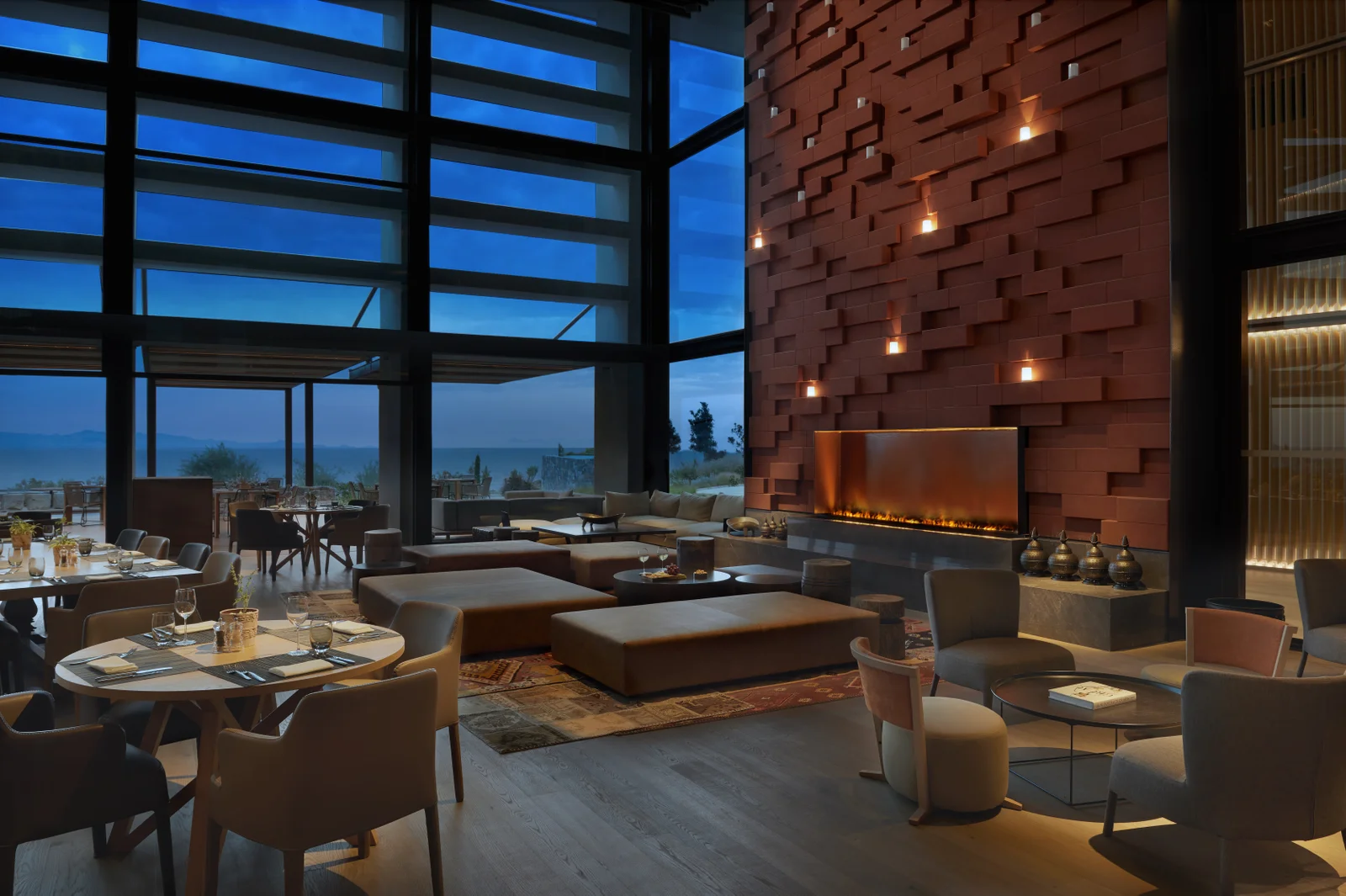 A double-height dining room at dusk, a long gas fire set into a sculpted terracotta wall and a wall of glass looking out to the sea.