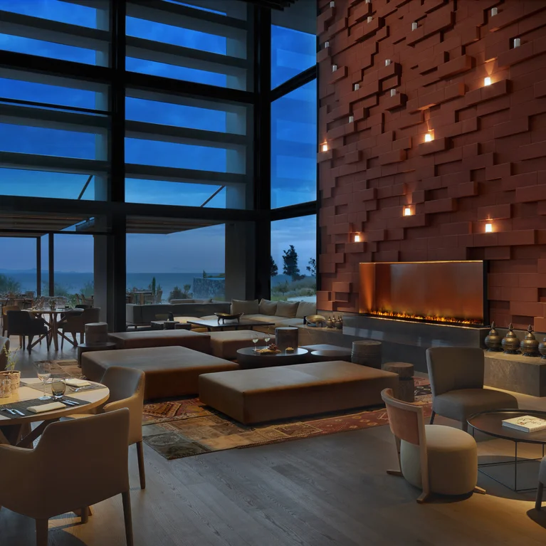 Un restaurante con materiales naturales, iluminación cálida, una acogedora chimenea y una amplia vista de la terraza y el paisaje nocturno gracias a los ventanales que van del suelo al techo.