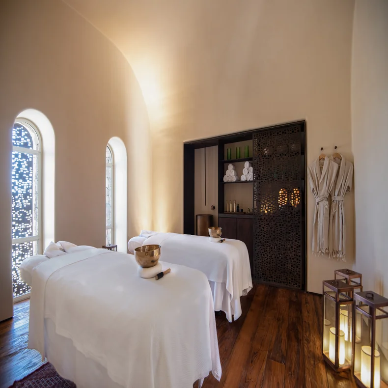 Spa suite featuring two massage tables with crisp linens, natural wood floors, gentle lighting, hanging robes, and neatly arranged towels and wellness items.