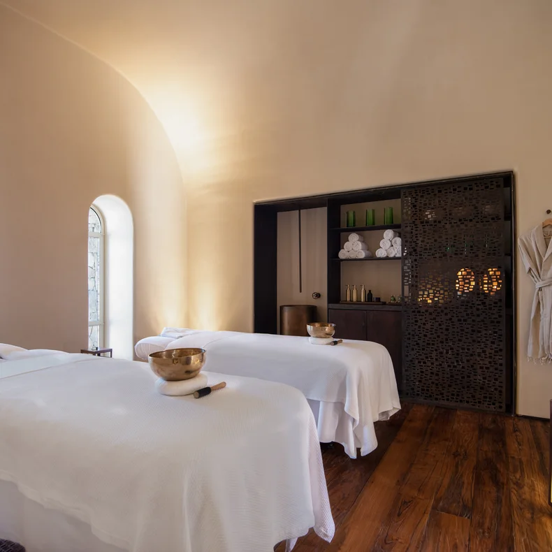 Spa suite featuring two massage tables with crisp linens, natural wood floors, gentle lighting, hanging robes, and neatly arranged towels and wellness items.