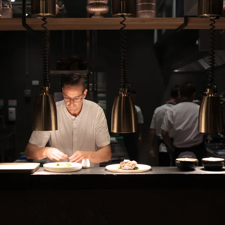 يقوم أحد محترفي الطهي بترتيب طبق تحت إضاءة خافتة في مطبخ مصمم بعناية ومنخفض الإضاءة. تصميم يسهل الوصول إليه ومواد طبيعية مرئية.