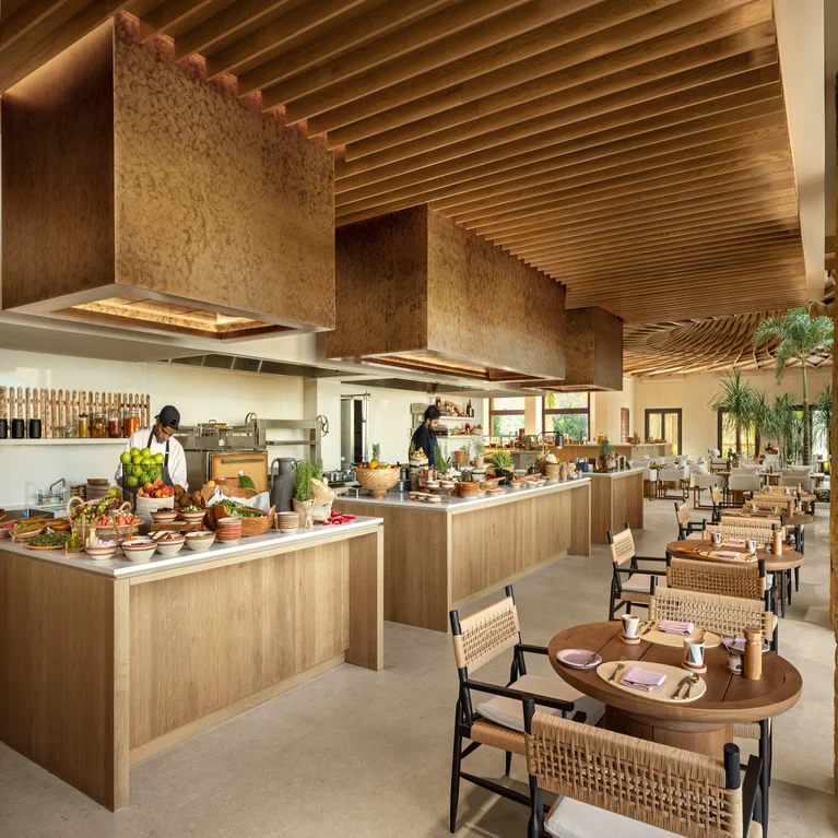 Open-plan restaurant featuring natural wood details, chefs at work, fresh ingredients, and thoughtfully arranged tables with woven seating.