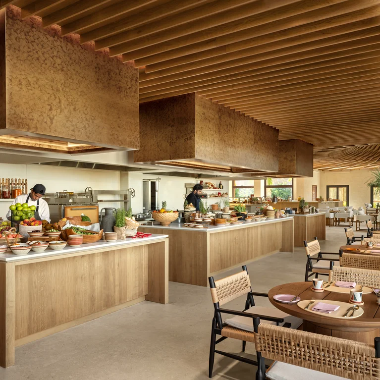 Open-plan restaurant featuring natural wood details, chefs at work, fresh ingredients, and thoughtfully arranged tables with woven seating.