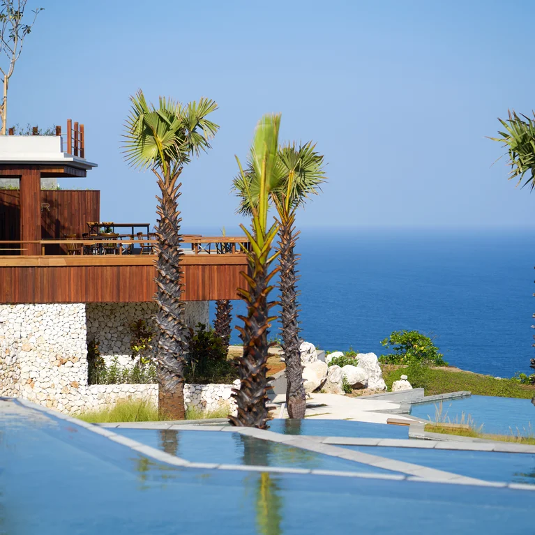 Villa sostenible de piedra natural y madera, bordeada de palmeras, con piscina infinita y vistas despejadas al mar bajo un cielo despejado.